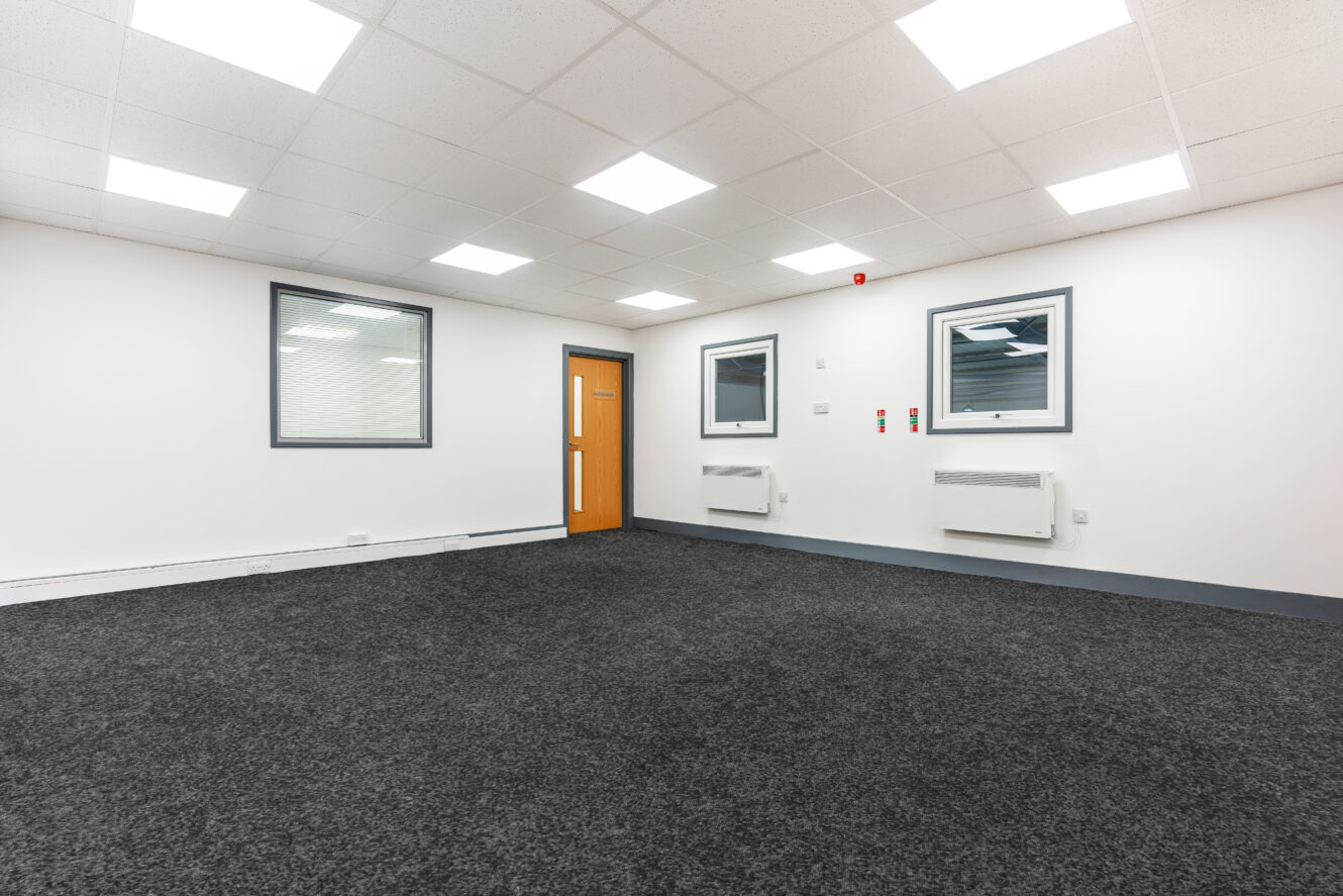 An empty office room with white walls, two windows, a wooden door, grey carpet, wall heaters, and bright ceiling lights.