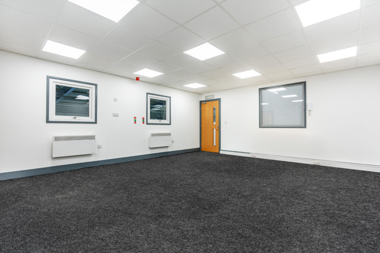 Empty office room with grey carpet, white walls, recessed ceiling lights, two windows, a wooden door, and wall-mounted heaters.