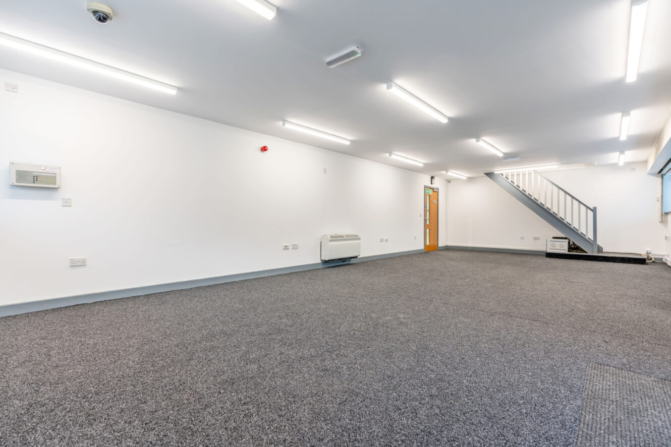 Empty office space with gray carpet, white walls, ceiling lights, a wall-mounted heater, and a metal staircase leading to an upper level.