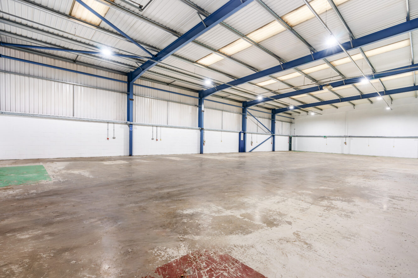 Interior of a large, empty warehouse with high ceilings, exposed beams, concrete floor, white walls, and fluorescent lighting.