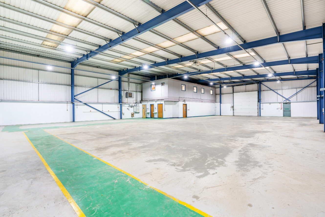 Interior of a large, empty industrial warehouse with high ceilings, overhead lights, and a small raised office area. The floor is marked with green and yellow lines.