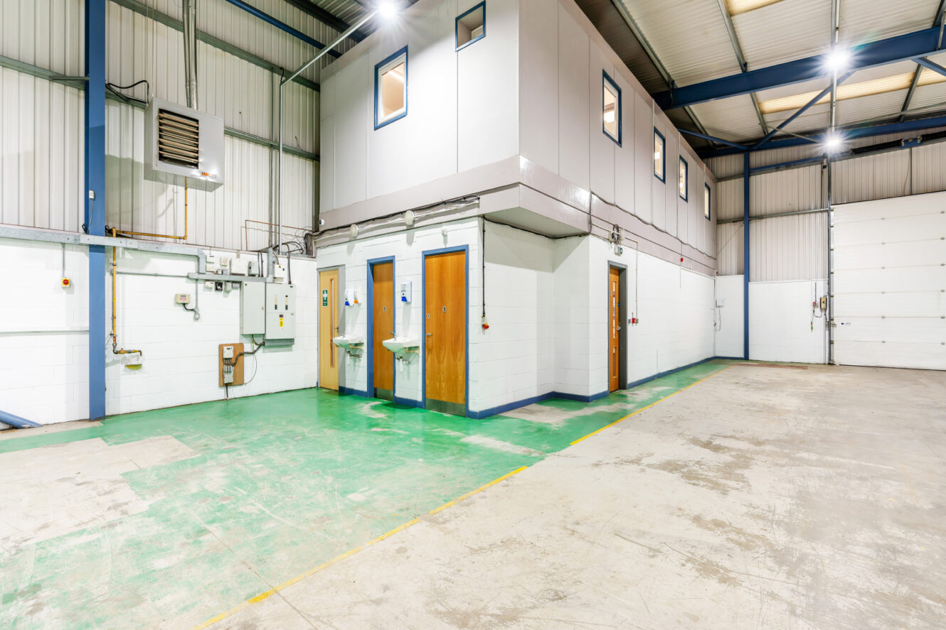 Interior of an empty industrial warehouse with a sectioned-off office area, multiple doors, sinks, green painted floor section, and white walls.