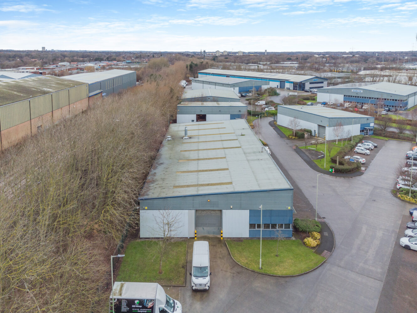 Aerial view of industrial warehouses with adjacent parking areas, vehicles, and surrounding trees under a partly cloudy sky.