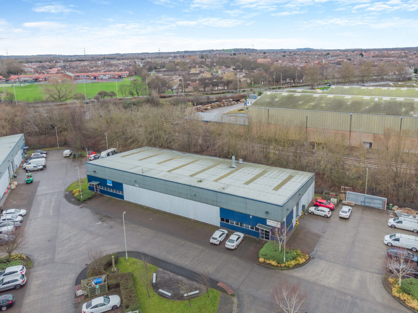 Aerial view of a large industrial warehouse with a parking lot, surrounded by trees and other buildings, with a sports field and residential area in the background.