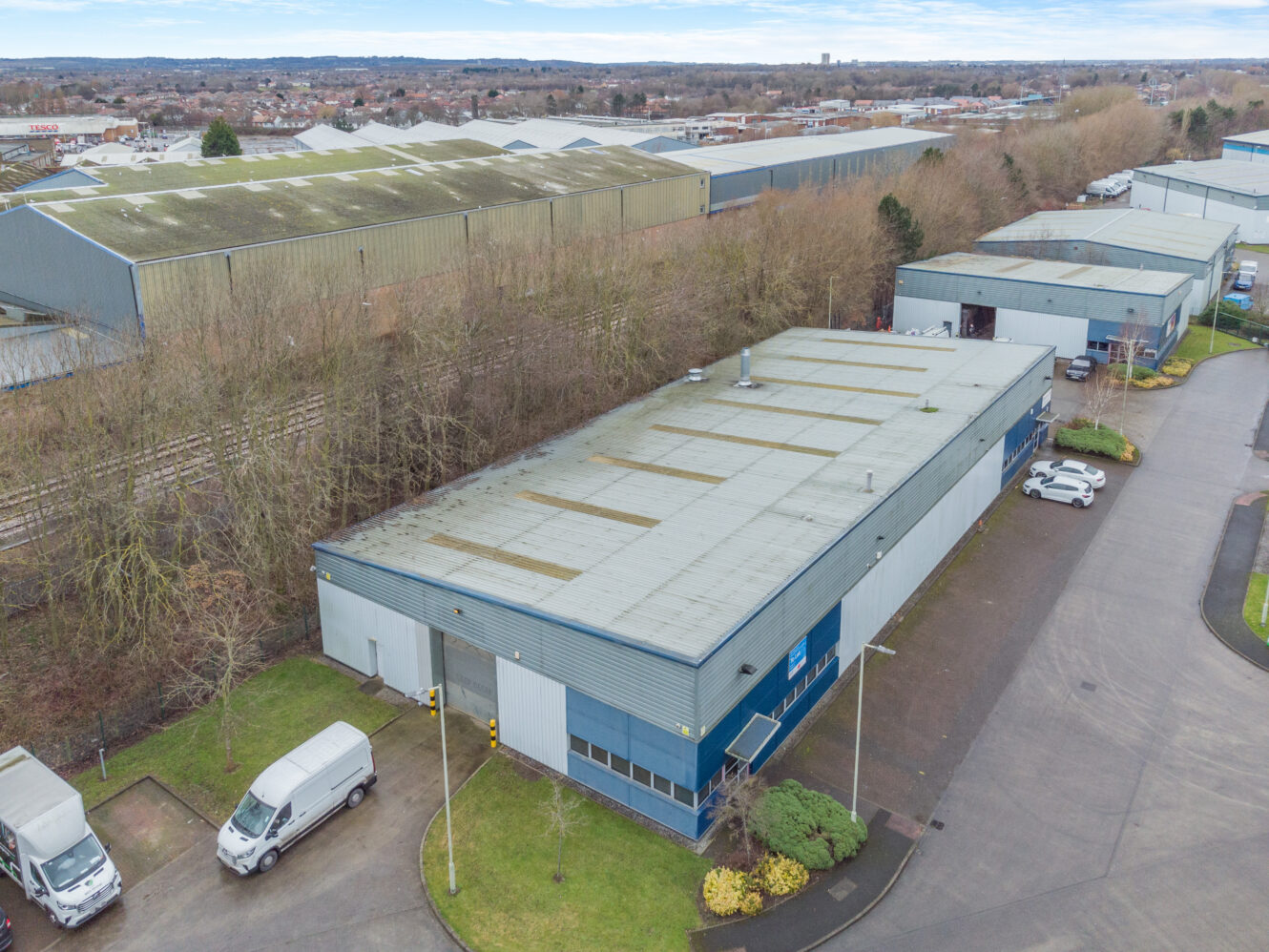Aerial view of a large industrial warehouse with a grey roof, surrounded by greenery, parked vans, and other industrial buildings in the background.