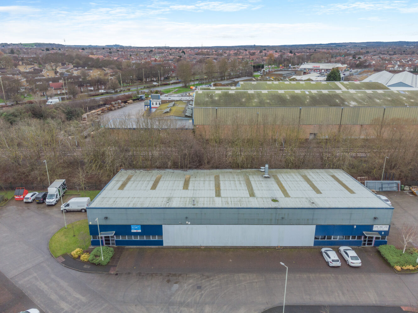 Aerial view of a blue and gray industrial warehouse with cars parked in front, surrounded by trees and neighboring industrial buildings.
