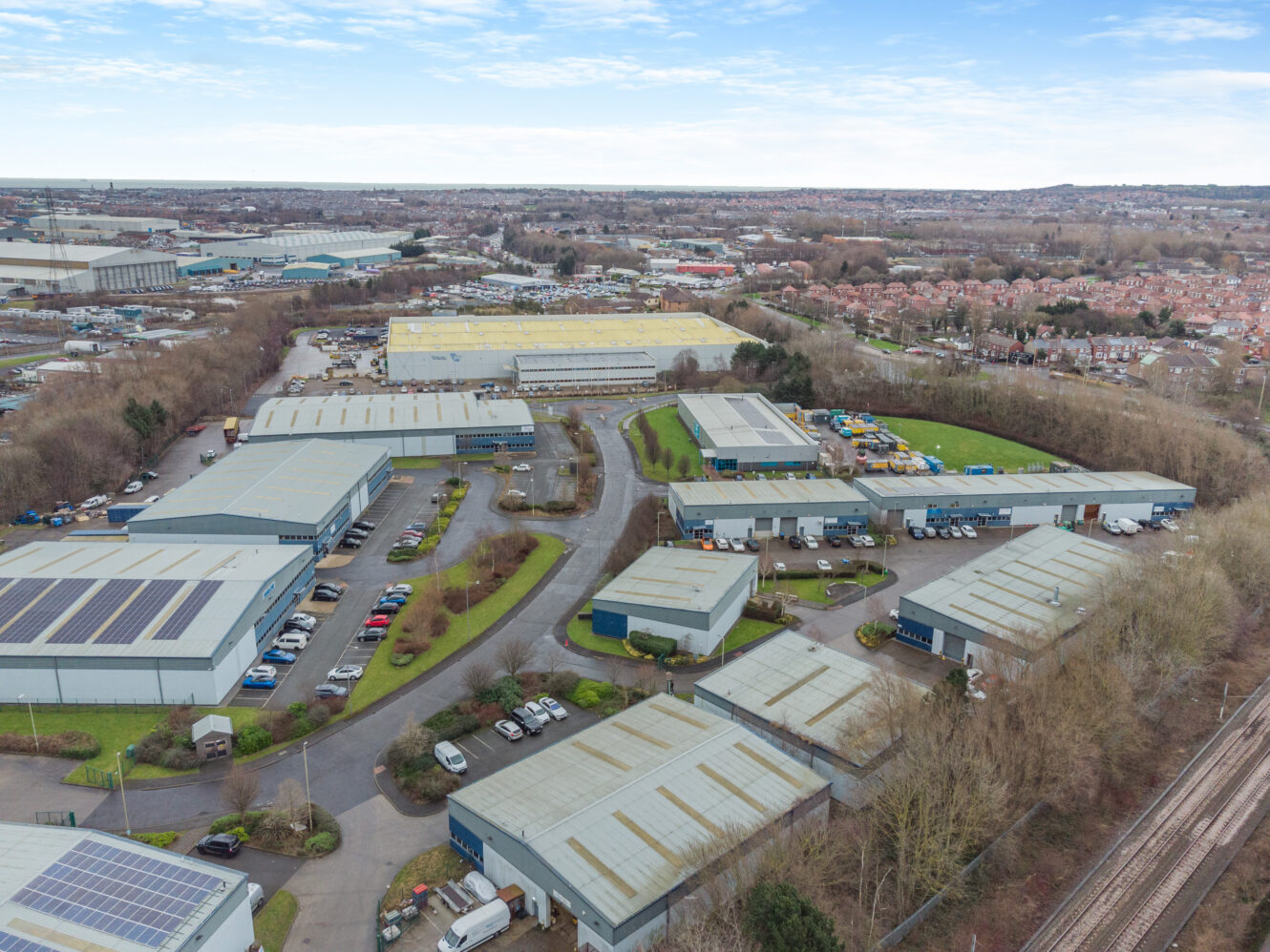 Aerial view of an industrial estate with multiple warehouses, parking lots, and surrounding trees, set near a residential area under a partly cloudy sky.