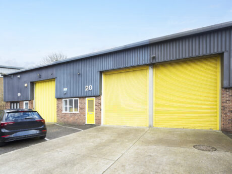 Industrial building with two large yellow roller doors, a yellow entrance door, unit numbers 19 and 20, brick lower walls, and a black car parked outside.