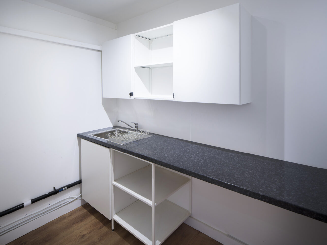 Minimalist kitchen with white cabinets, black countertop, stainless steel sink, and open shelving. The space is clean and unfurnished with wood flooring.