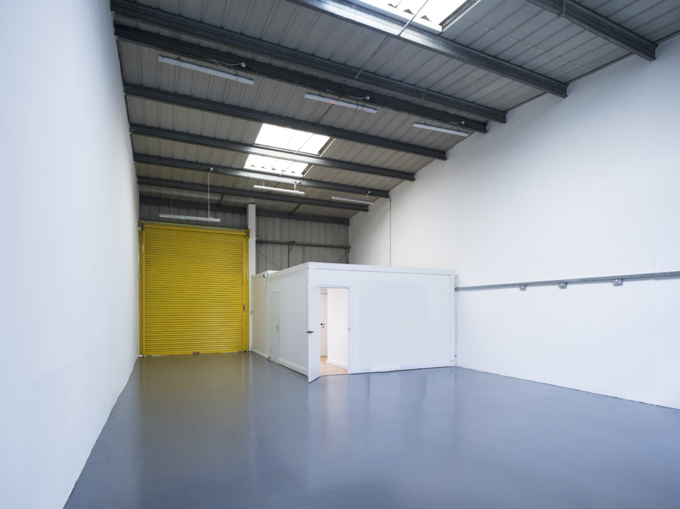 Empty industrial warehouse with a gray floor, white walls, a small white office unit, and a large yellow roller door at the end. Natural light enters through ceiling windows.