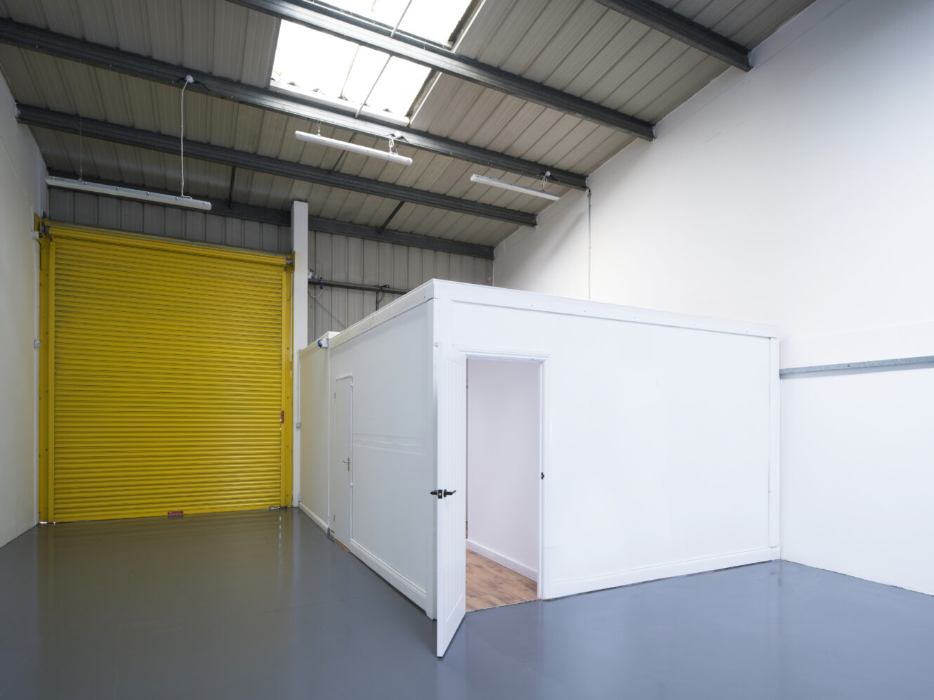 A clean industrial space with a white interior office structure, an open door, gray floor, and a large yellow roller shutter door.