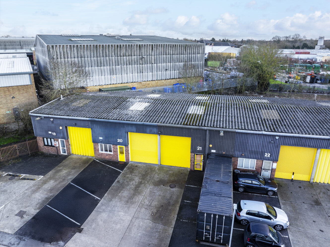 Aerial view of industrial units with gray roofs and bright yellow doors, with several parked cars and a shipping container in the foreground.