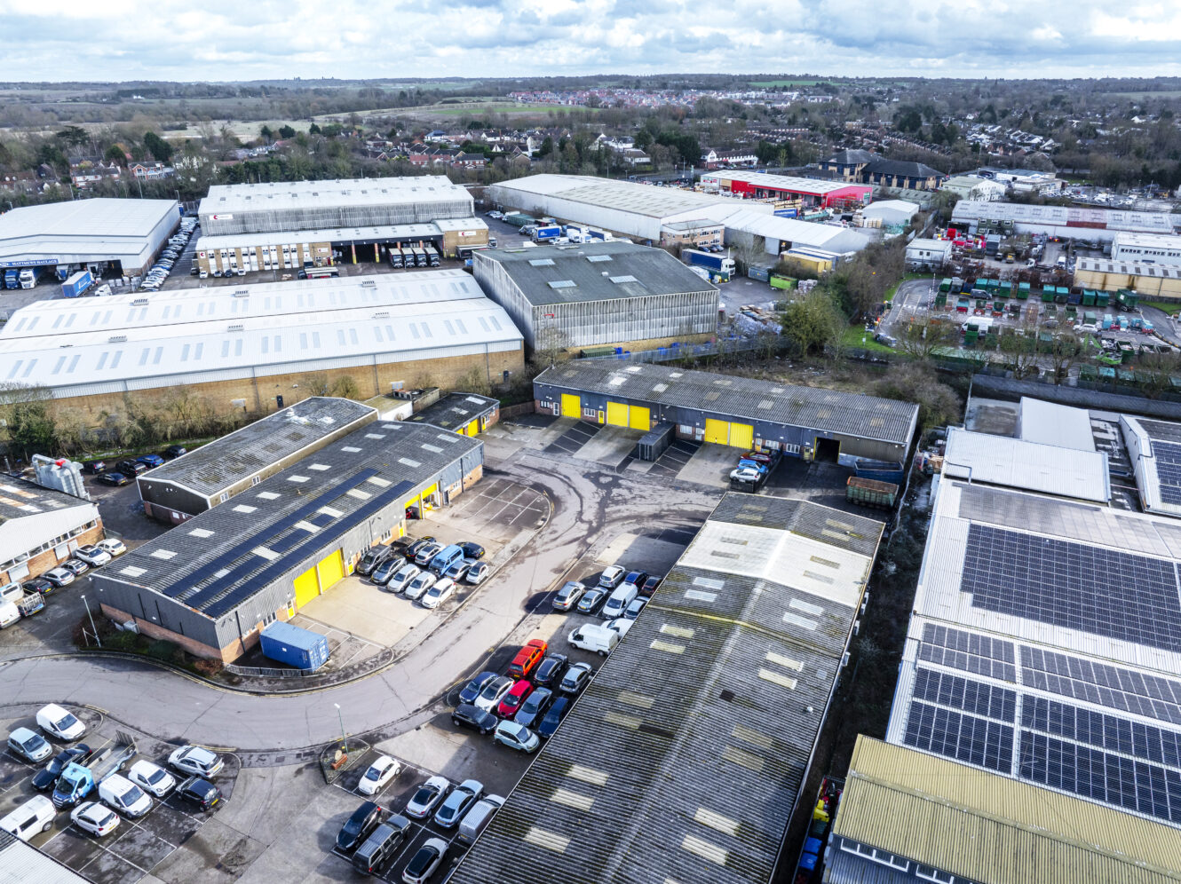 Aerial view of an industrial estate with several warehouses, parked cars, and surrounding roads under a cloudy sky.