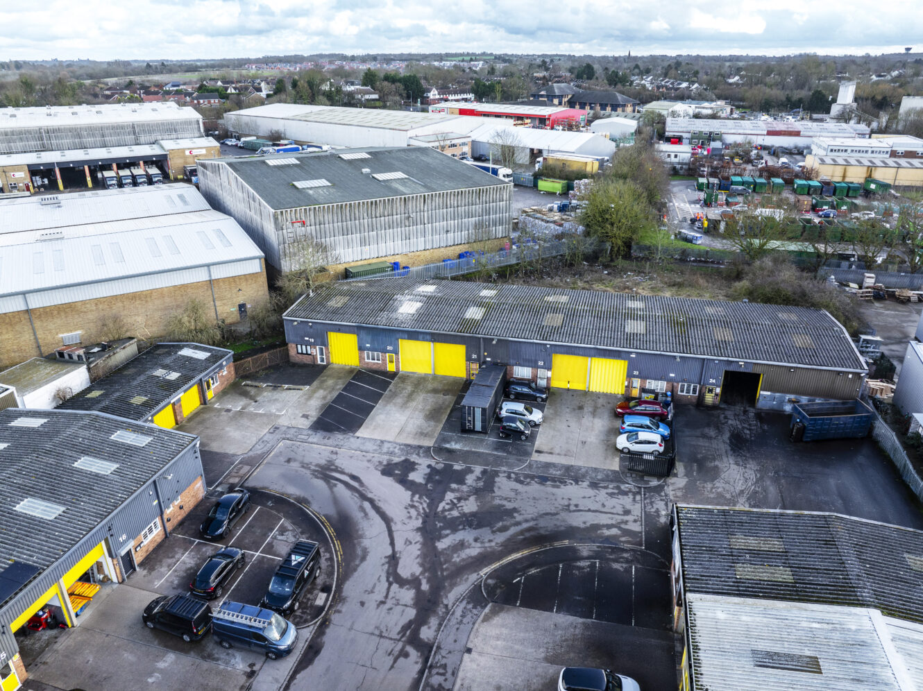 Aerial view of an industrial estate with warehouses, parked vehicles, and surrounding roads on a cloudy day.