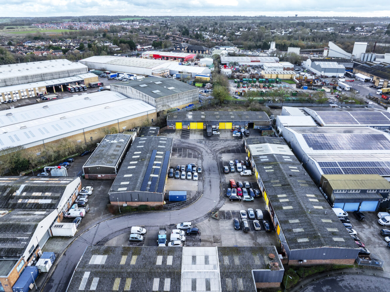 Aerial view of an industrial estate with multiple warehouses, parked cars, and surrounding roads, set in a suburban area with fields and residential buildings in the background.