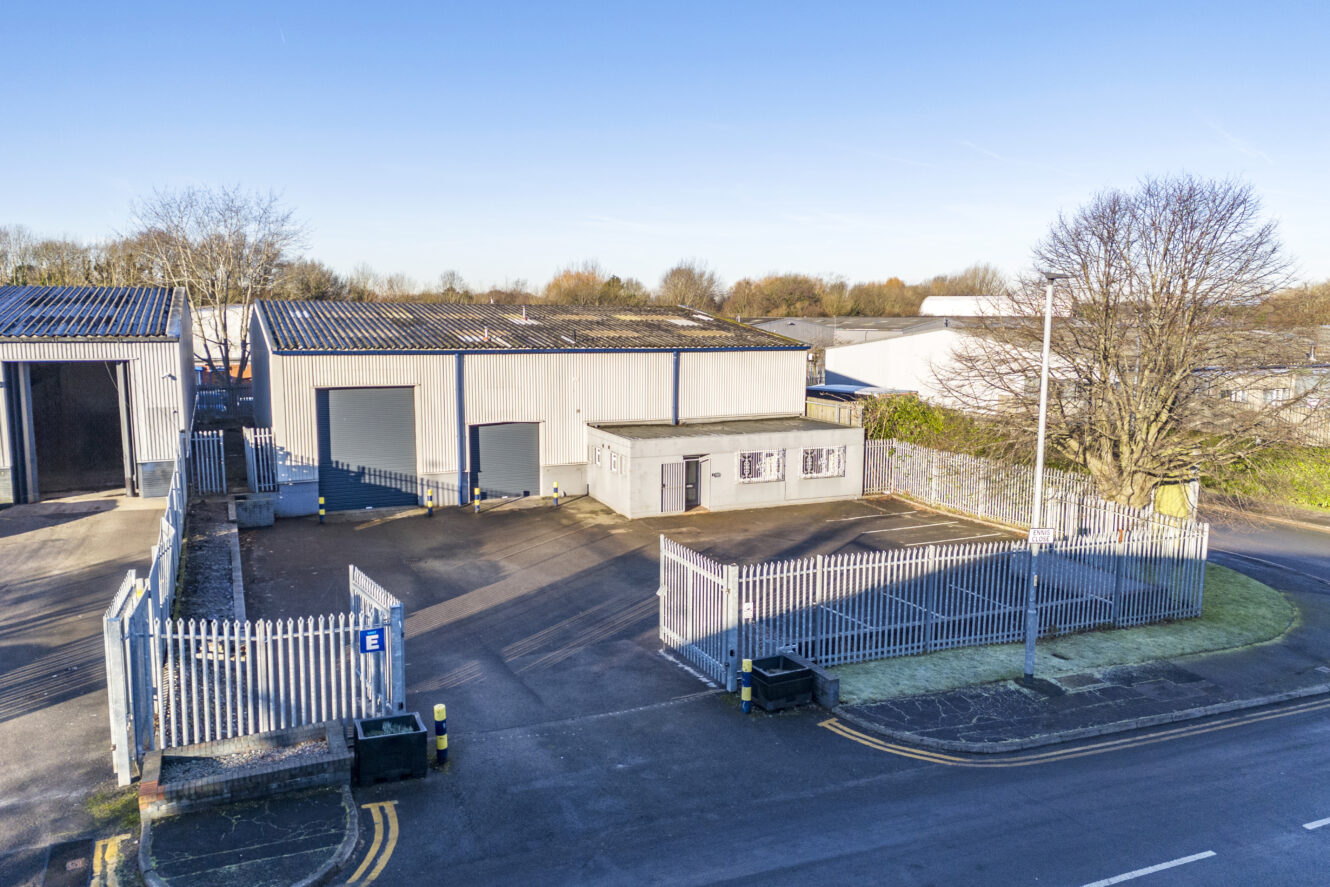 A fenced industrial facility with a gray warehouse, large roller doors, a small office building, and a gated entrance beside a road on a clear day.
