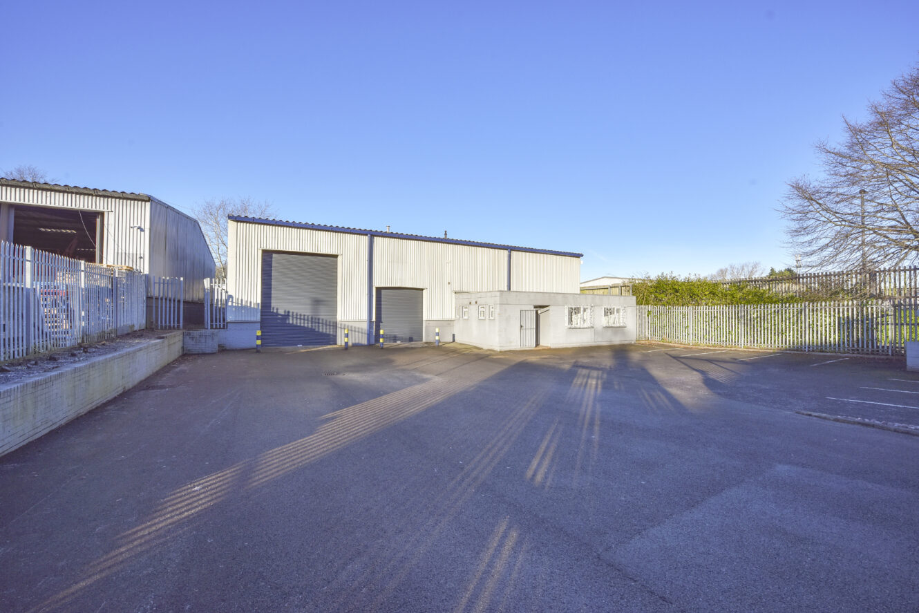 A large industrial warehouse with two wide garage doors and fenced surroundings, set on a paved lot under a clear blue sky.