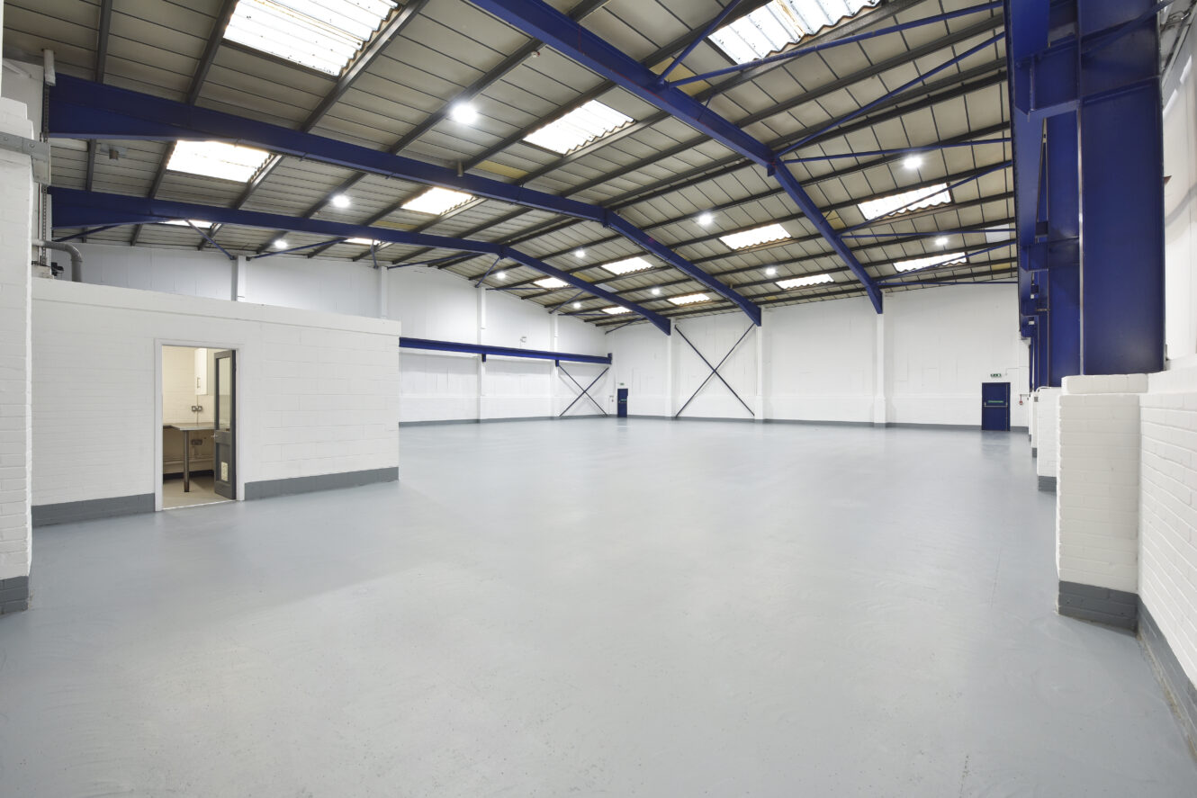 Interior of a large, empty industrial warehouse with high ceilings, exposed beams, white walls, and a small room with a sink in one corner.