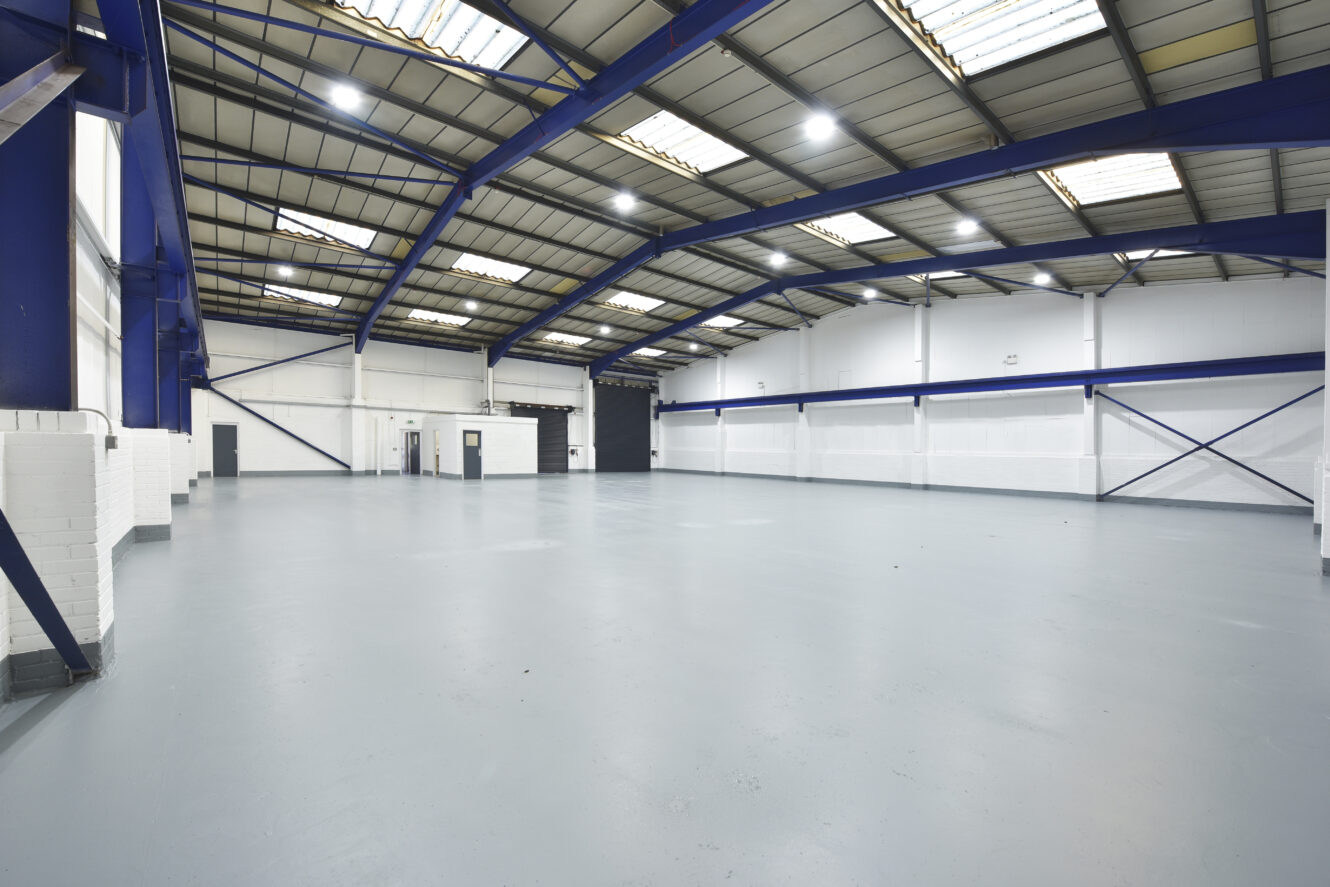 Large, empty industrial warehouse with high ceilings, white walls, blue steel beams, and polished grey floor, illuminated by overhead fluorescent lights.