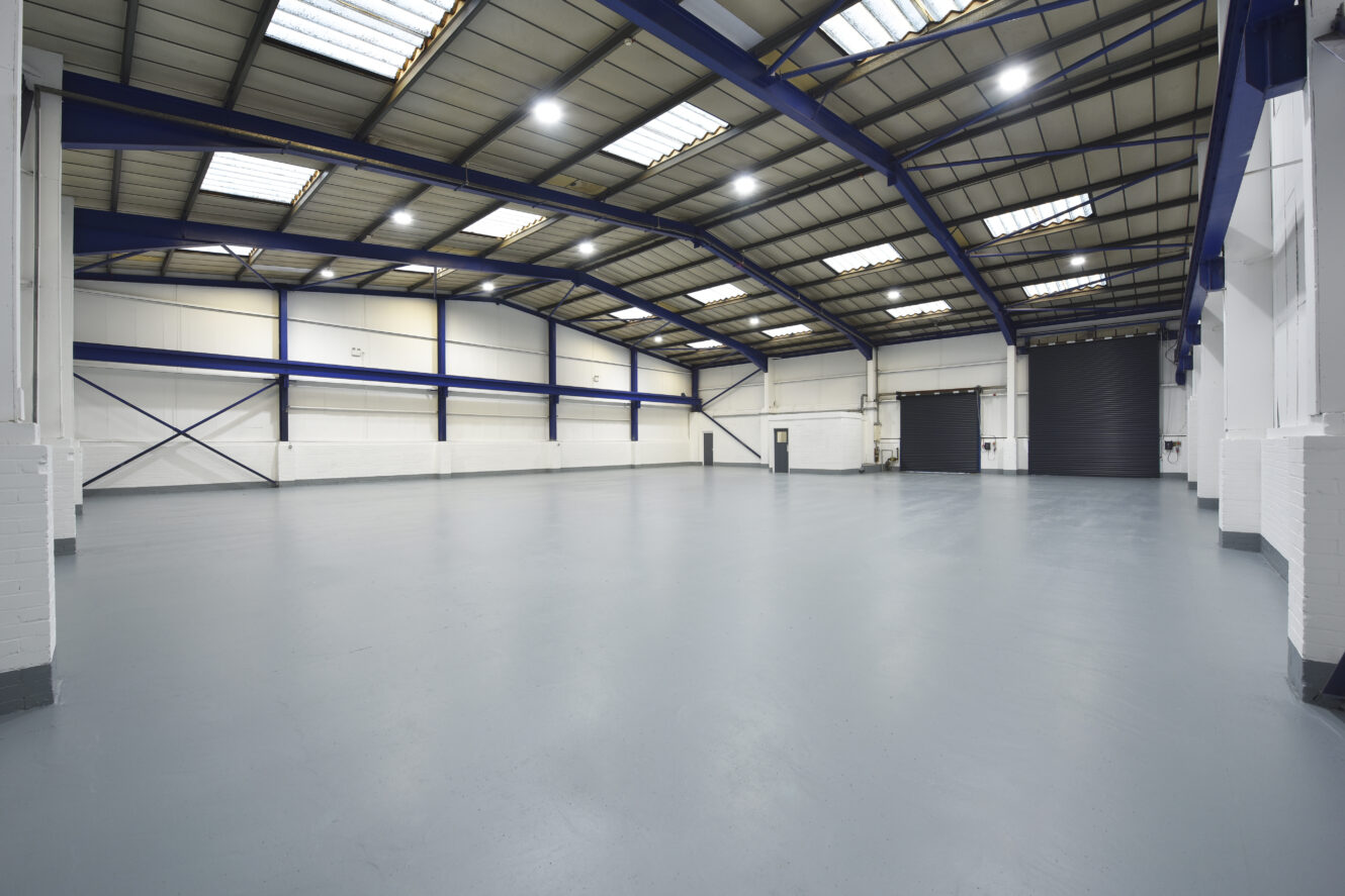Spacious, empty warehouse with high ceilings, skylights, white walls, and a large closed roller shutter door.
