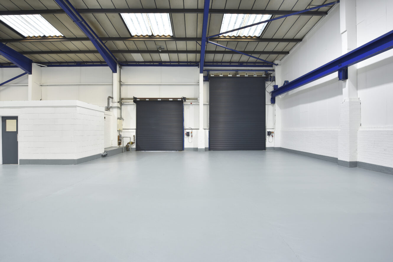 Empty industrial warehouse with a smooth light gray floor, white walls, high ceiling, blue beams, and two large black roller shutter doors.