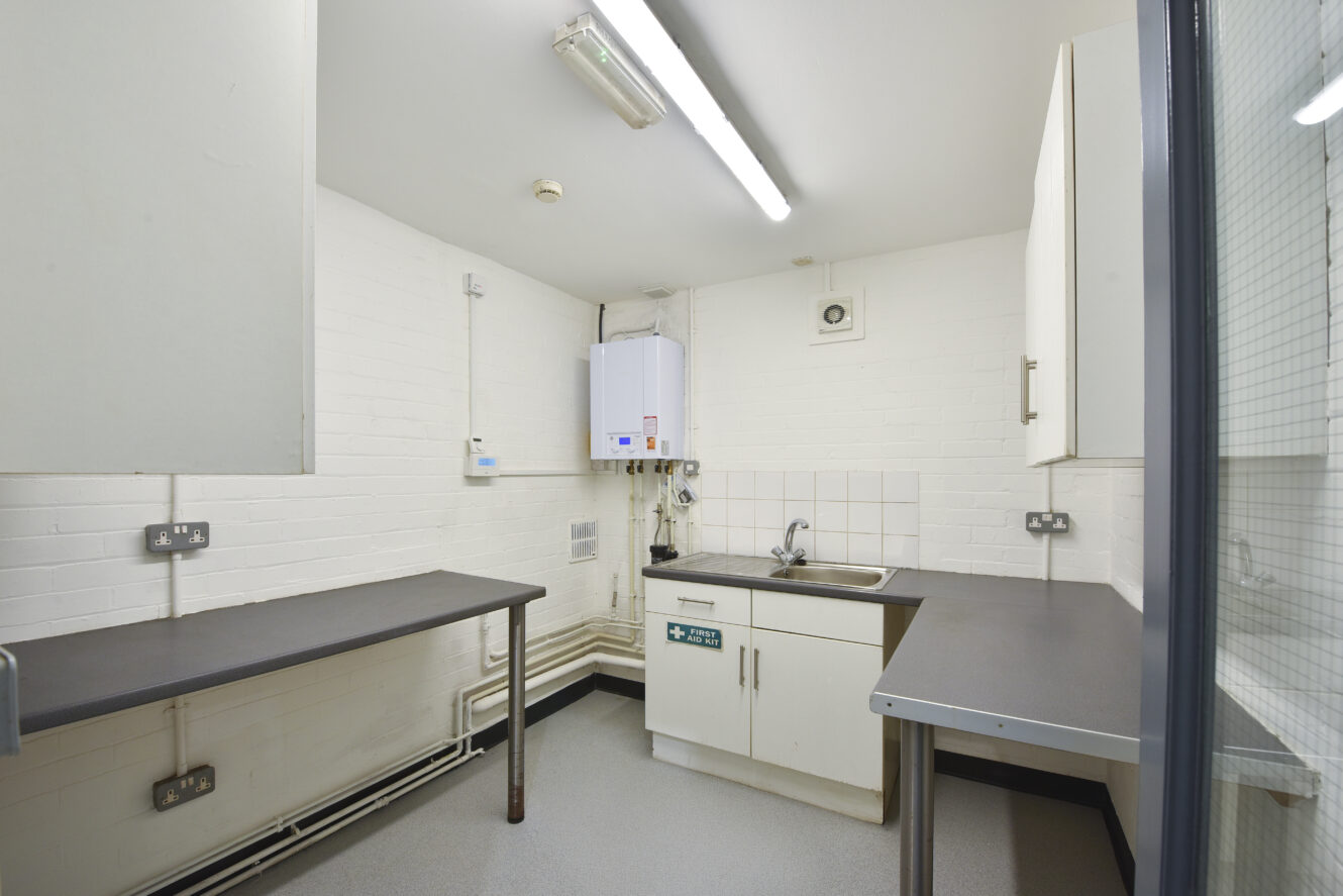 Small utility room with white brick walls, grey countertops, a sink, wall-mounted boiler, overhead fluorescent lights, and minimal furnishings.