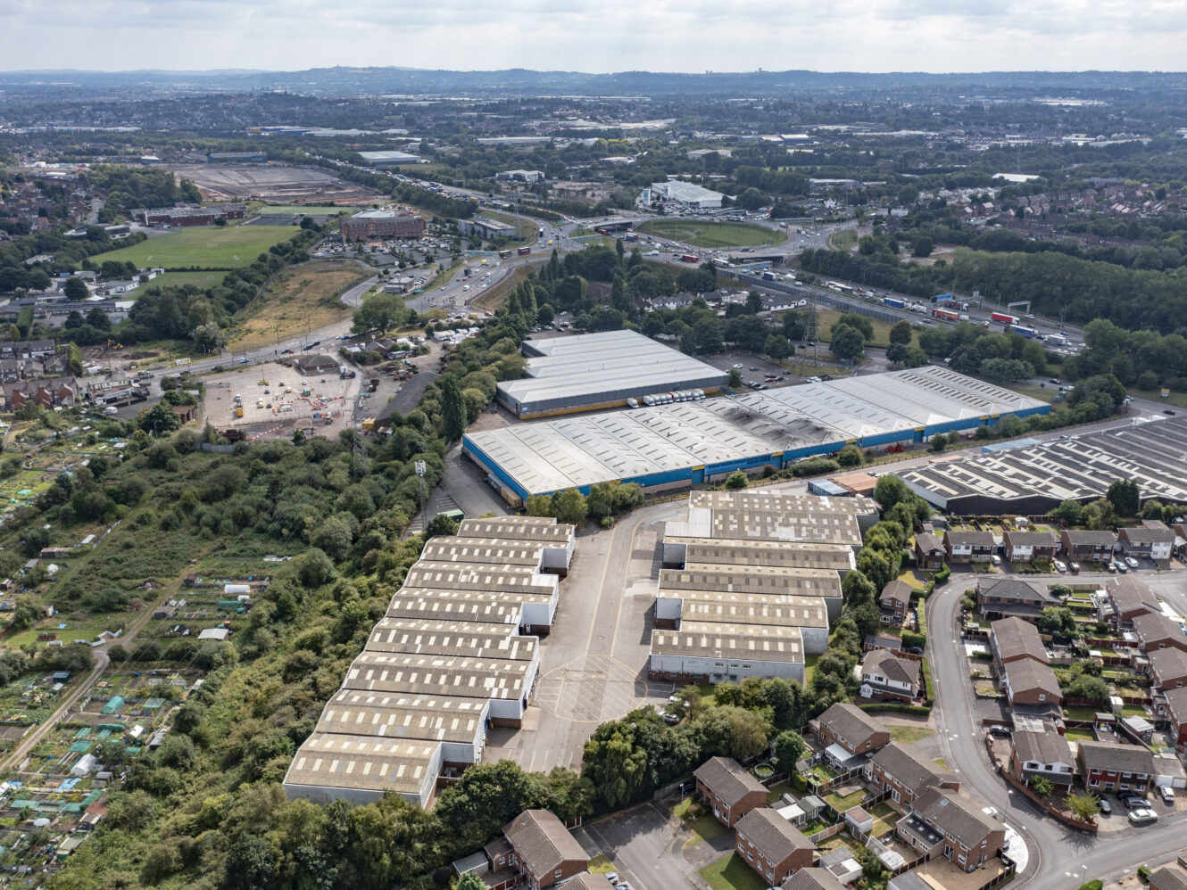Aerial view of an industrial estate with warehouses, nearby residential houses, allotments, and a major road with traffic in a suburban area.