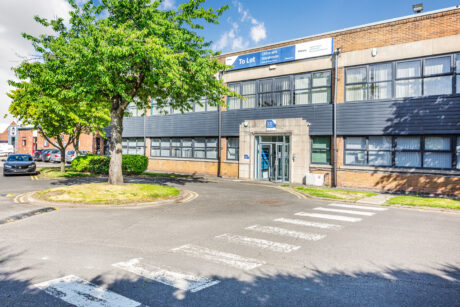 Two-story office building with To Let signs, large windows, a main entrance, and a tree by the roadside on a sunny day.