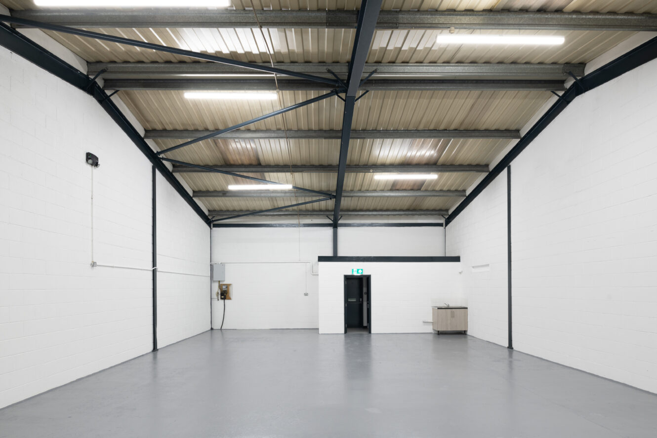 An empty industrial warehouse with white walls, a high metal ceiling, concrete floor, a small door, and a kitchenette in the back corner.