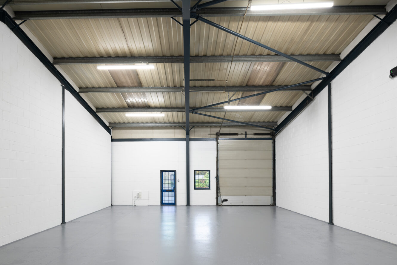 Empty industrial warehouse with white brick walls, grey concrete floor, exposed steel beams, a large roller door, and a small door with a window.