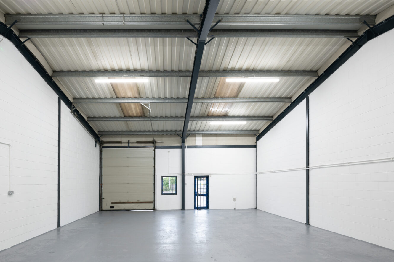 Empty industrial warehouse with white walls, concrete floor, high ceiling, overhead fluorescent lights, a large roller door, and a single entry door with a small window.