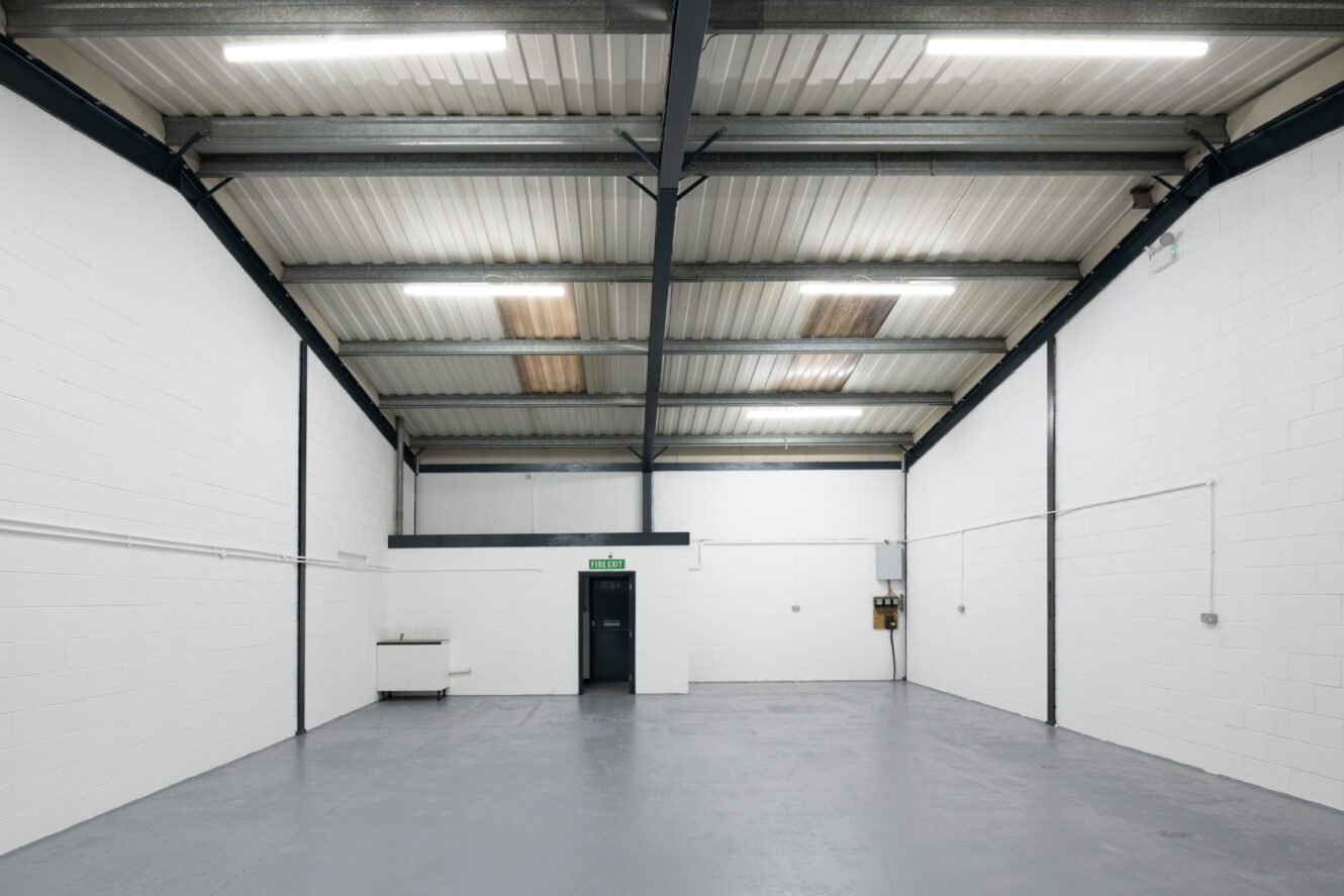 Empty industrial warehouse interior with white painted brick walls, grey concrete floor, metal ceiling beams, fluorescent lights, and a closed door at the far end.