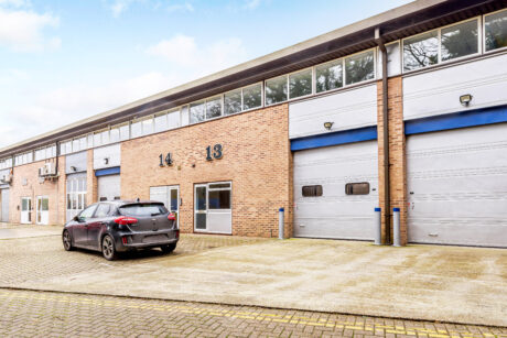 A black car is parked outside industrial units numbered 13 and 14, featuring large garage doors and brick exteriors.