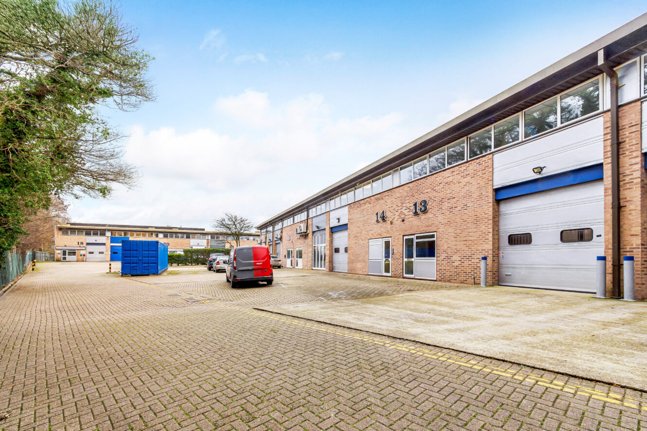 A small industrial estate with brick warehouse units, numbered 13 and 14, a red van, blue recycling container, and clear sky above.