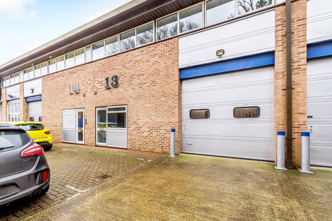 Brick commercial units with large windows and numbered doors, featuring parked cars and a concrete driveway in front.