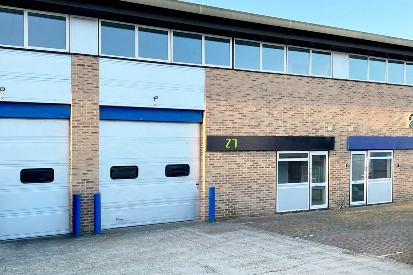A commercial building with brown brick walls, two white garage doors, and a glass entrance marked with the number 27.