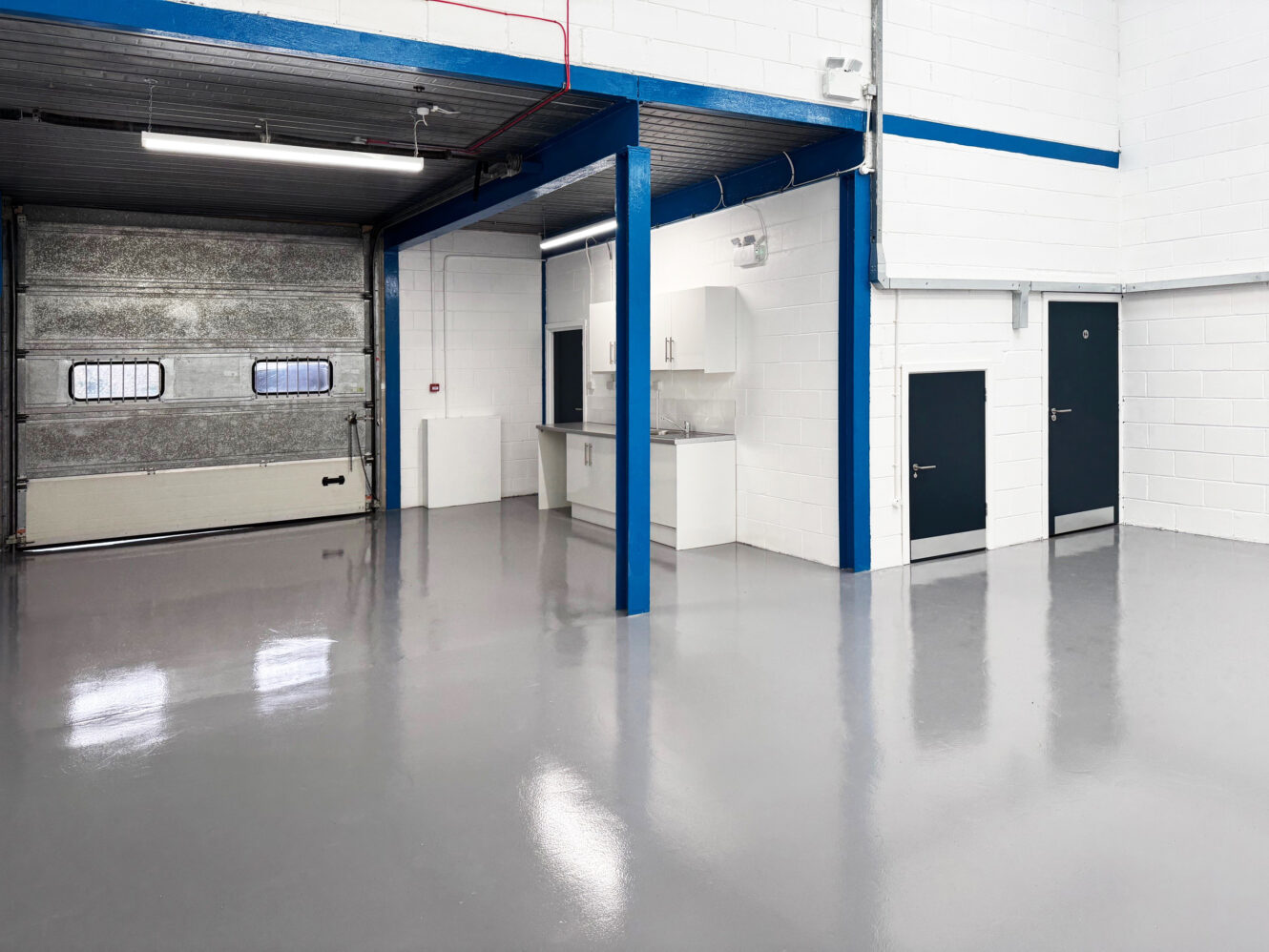 A clean, empty industrial warehouse with a polished concrete floor, a large metal garage door, blue structural beams, and a small kitchenette area.
