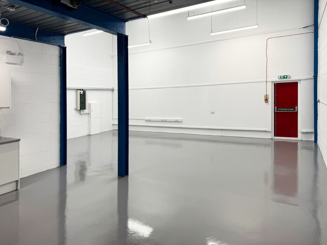 An empty industrial unit with white walls, blue support columns, a polished gray floor, fluorescent lighting, and a red emergency exit door.