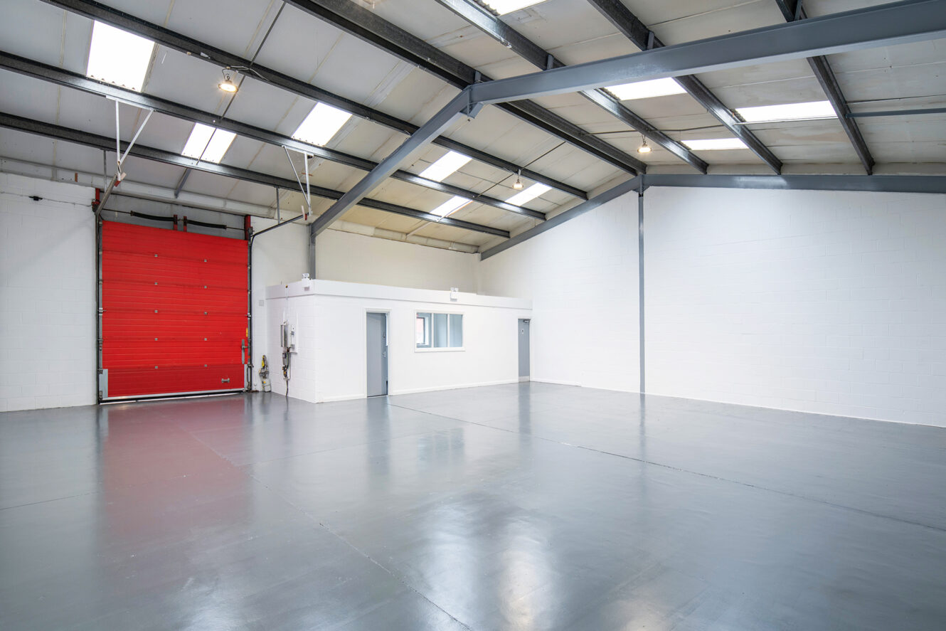 Empty warehouse interior with a polished concrete floor, white walls, a small office space, and a large red industrial door.