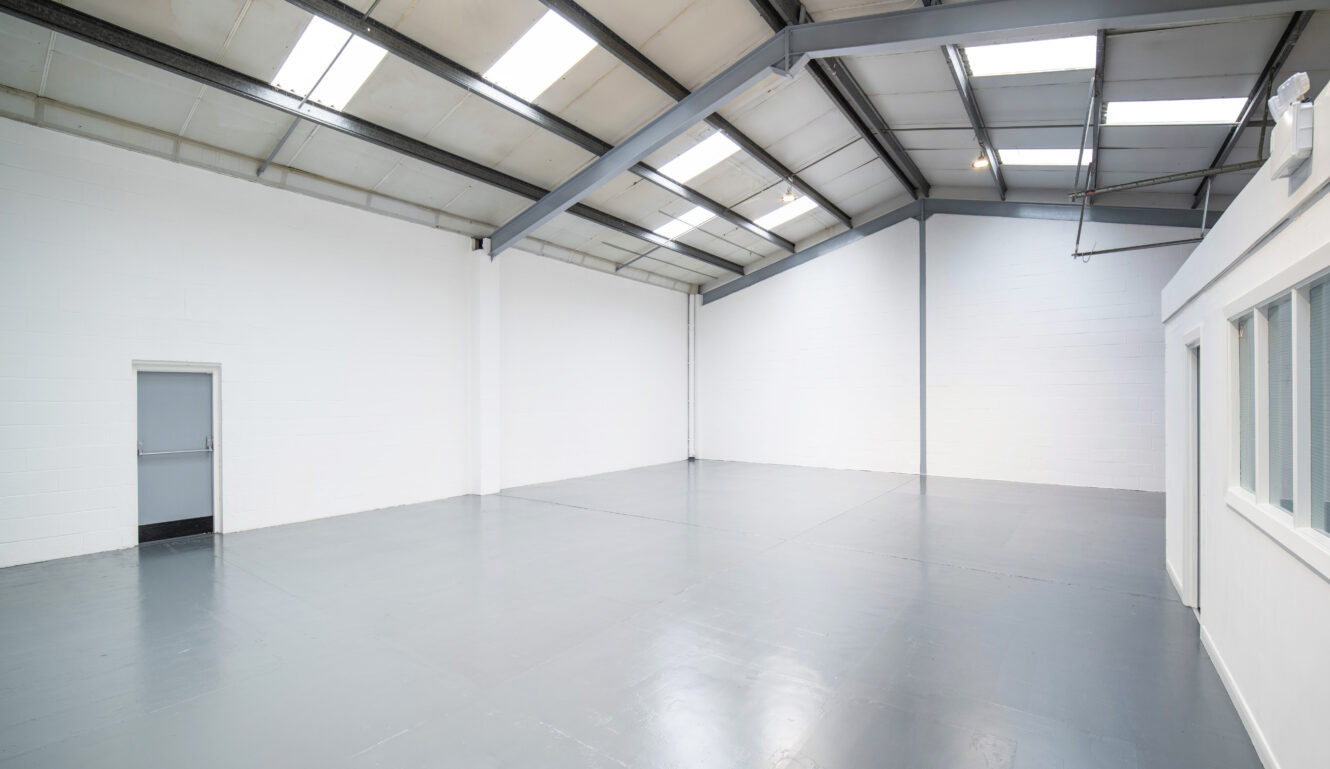 Large empty industrial warehouse with white walls, a high ceiling with skylights, a closed gray door, and a small office section with windows on the right.