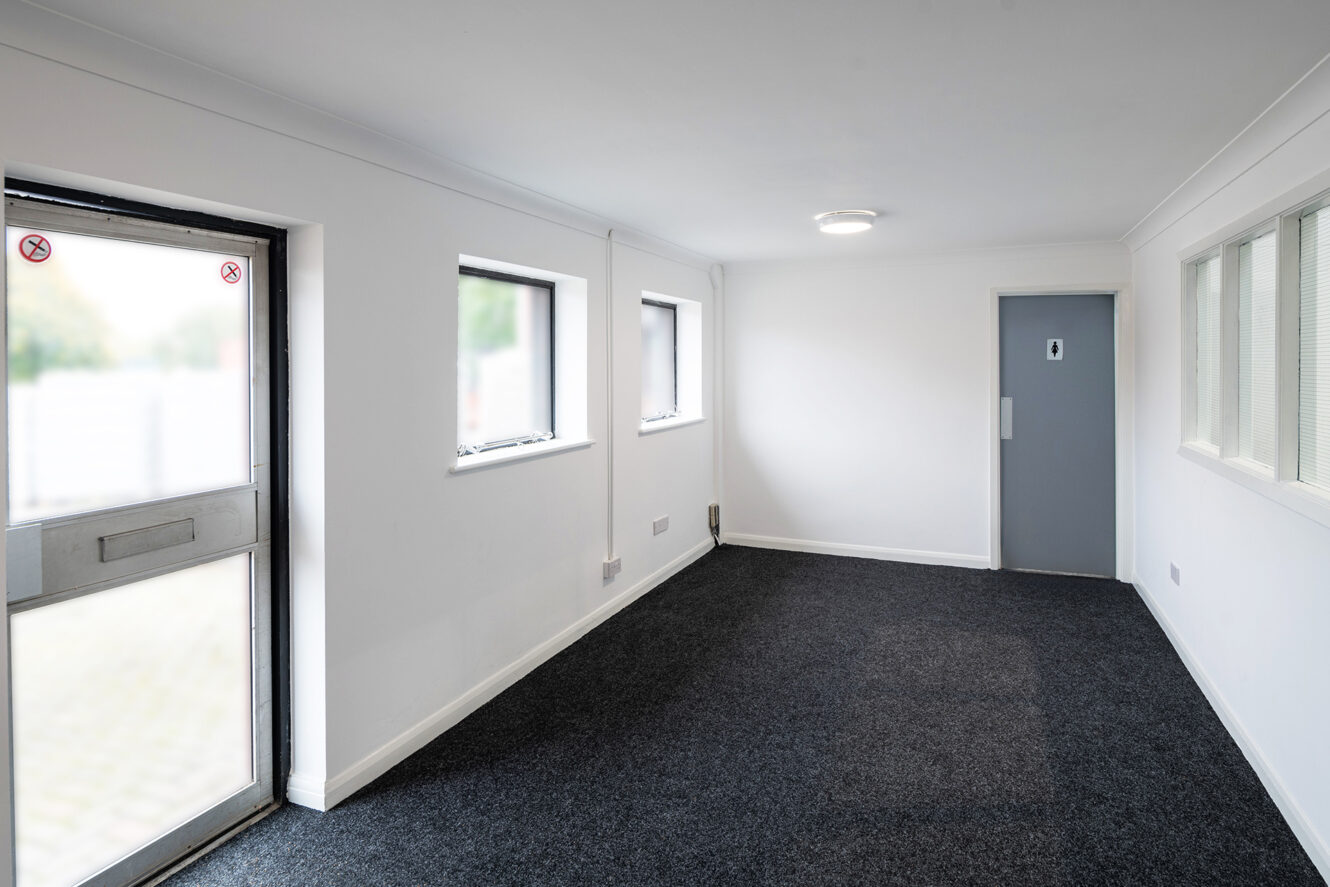 Empty room with white walls, three windows, a grey carpet, a closed grey door, and a glass exit door on the left. Ceiling light is on.
