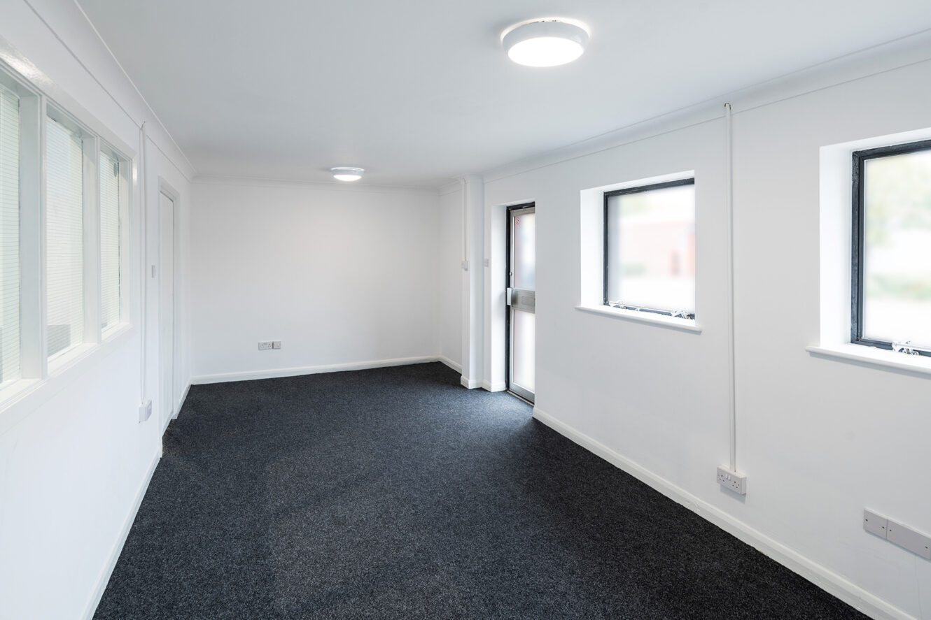Empty room with white walls, two windows, a door with frosted glass, and dark carpeted flooring under ceiling lights.