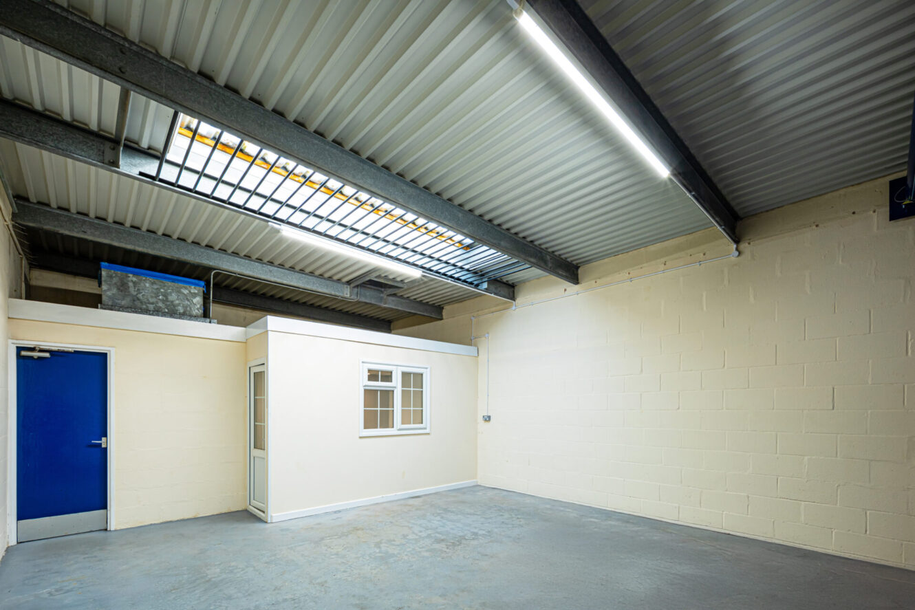 Empty industrial room with a concrete floor, cream-colored brick walls, a blue door, ceiling lights, and a small enclosed office with a window inside the space.