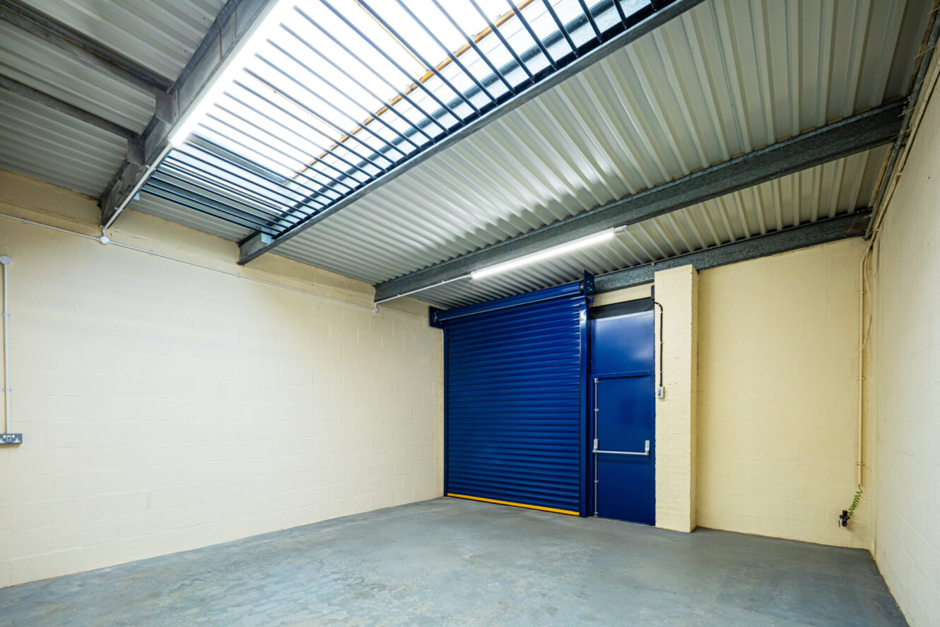 Empty industrial unit with cream walls, gray concrete floor, exposed ceiling beams, and a closed blue roller shutter door next to a matching blue personnel door. Fluorescent lighting overhead.