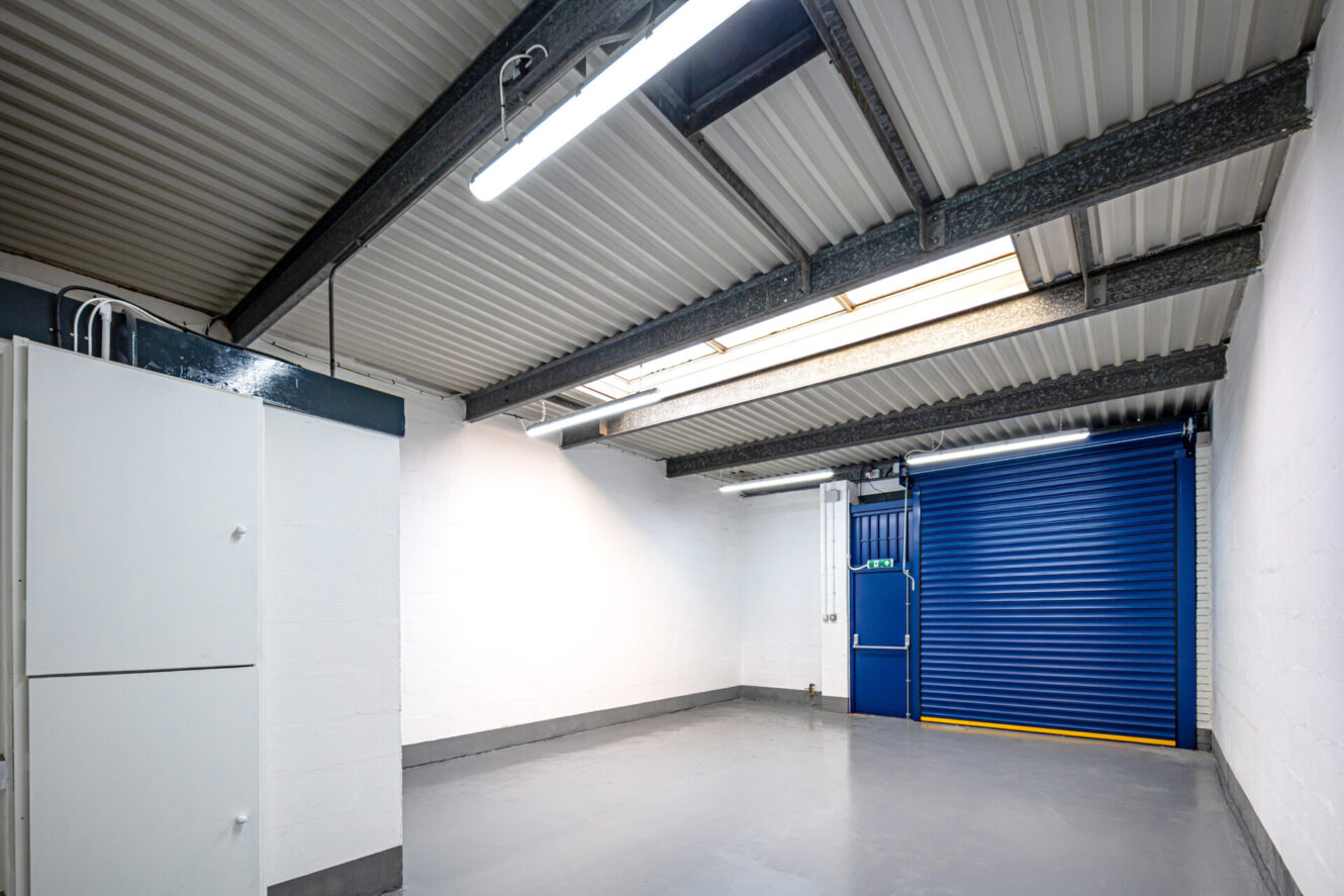 A clean, empty garage with white walls, grey floor, overhead lights, metal ceiling beams, a blue roller shutter door, and white storage cabinets.