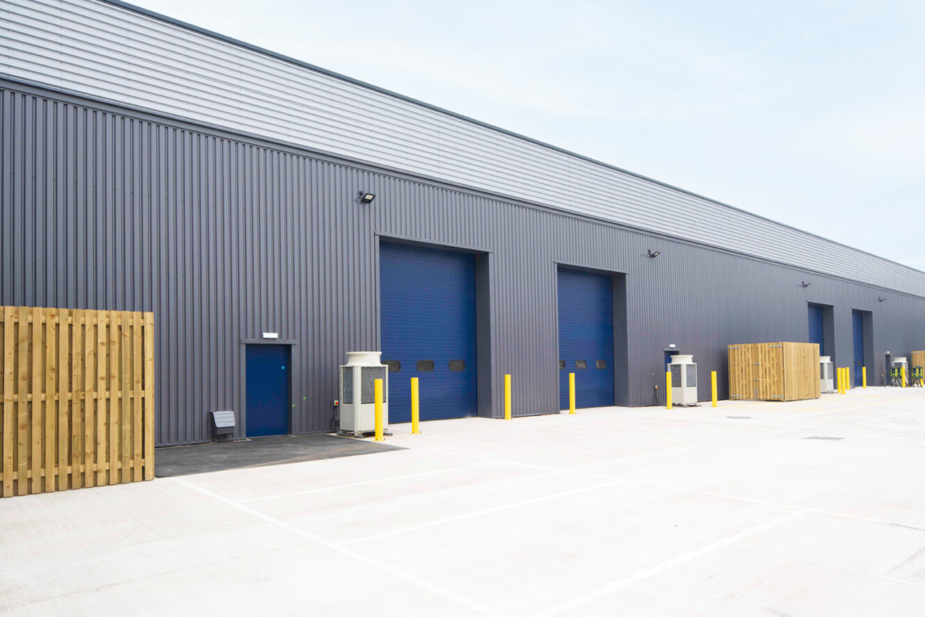 A modern industrial warehouse exterior with gray metal siding, blue doors, multiple loading bays, and yellow bollards in a spacious paved lot.