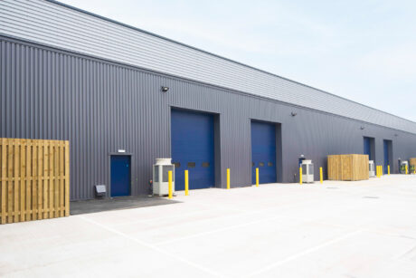 A modern industrial warehouse exterior with gray metal siding, blue doors, multiple loading bays, and yellow bollards in a spacious paved lot.