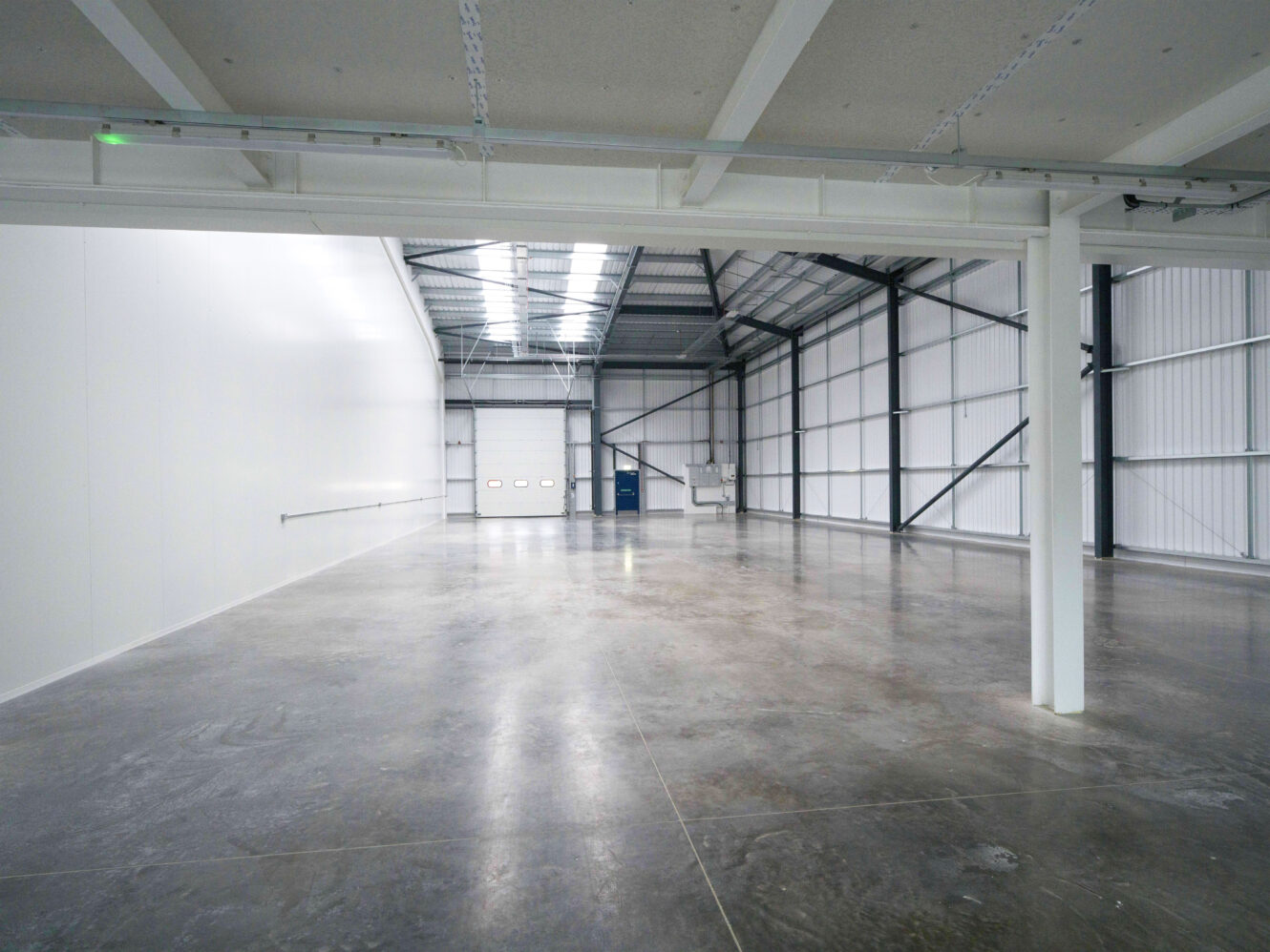 Empty industrial warehouse with polished concrete floors, high ceilings, white walls, and a large roll-up door at the far end.