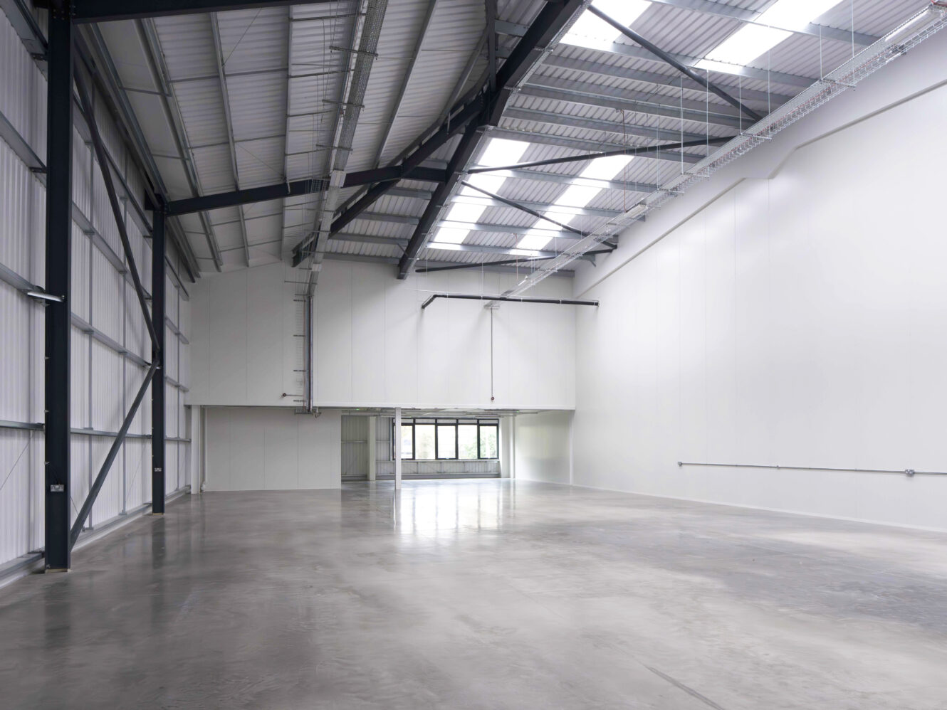 Interior of an empty, modern industrial warehouse with polished concrete floors, white walls, large windows, and exposed steel beams.