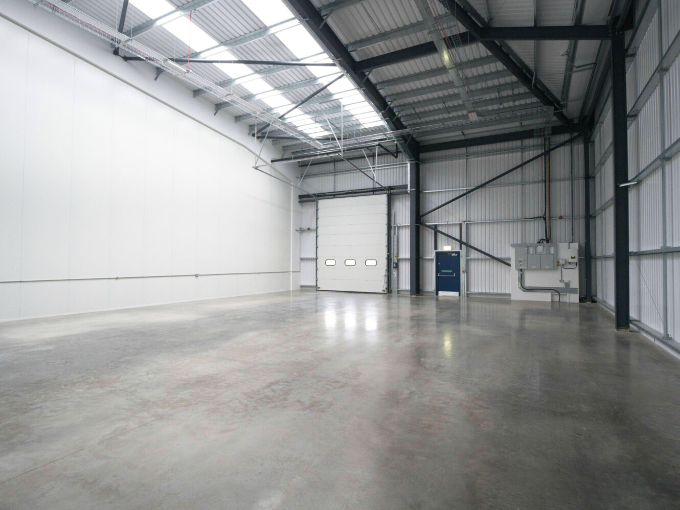 Interior of an empty industrial warehouse with polished concrete floor, high ceiling, metal beams, large white wall, roller shutter door, and a blue access door.